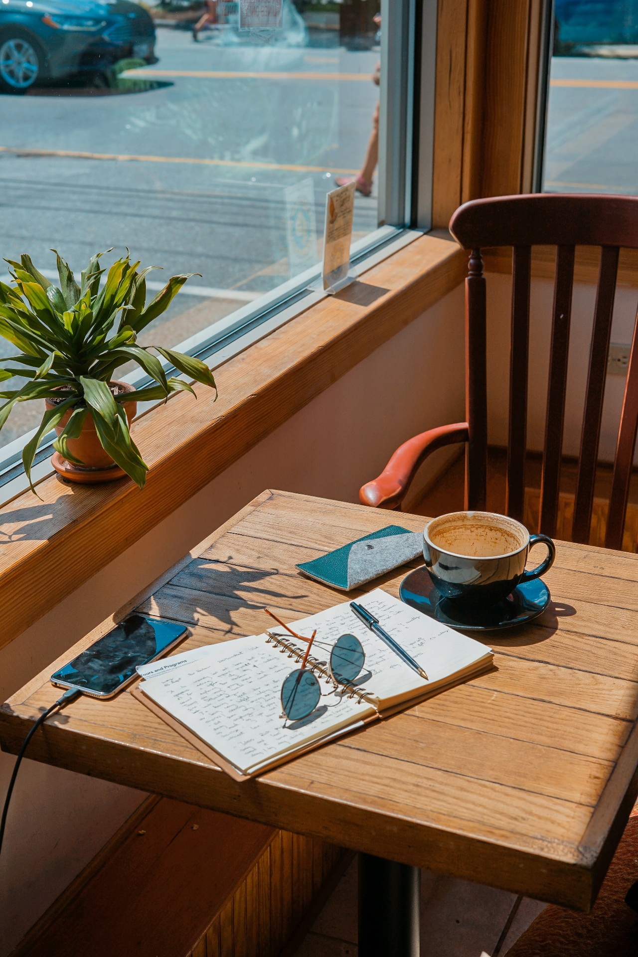 Open journal, sunglasses, and a coffee cup on a wooden table by a sunny window — calm morning