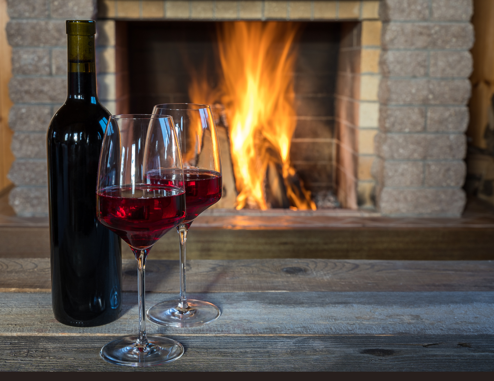 A bottle of red wine and two glasses on a wooden mantle in front of a glowing fireplace